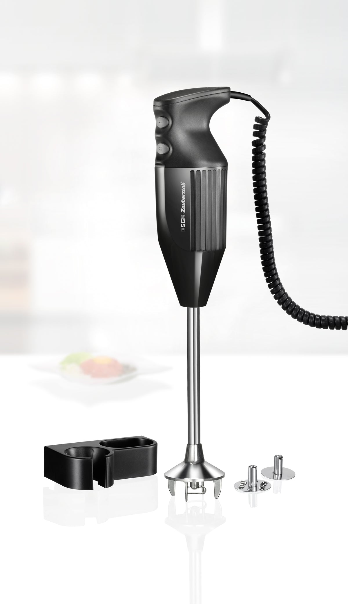Ein schwarzer Stabmixer mit Spiralkabel steht aufrecht auf einer weißen Fläche, begleitet von einer Wandhalterung und drei metallischen Mixaufsätzen. Im Hintergrund ist eine unscharfe Küchenszene zu sehen.