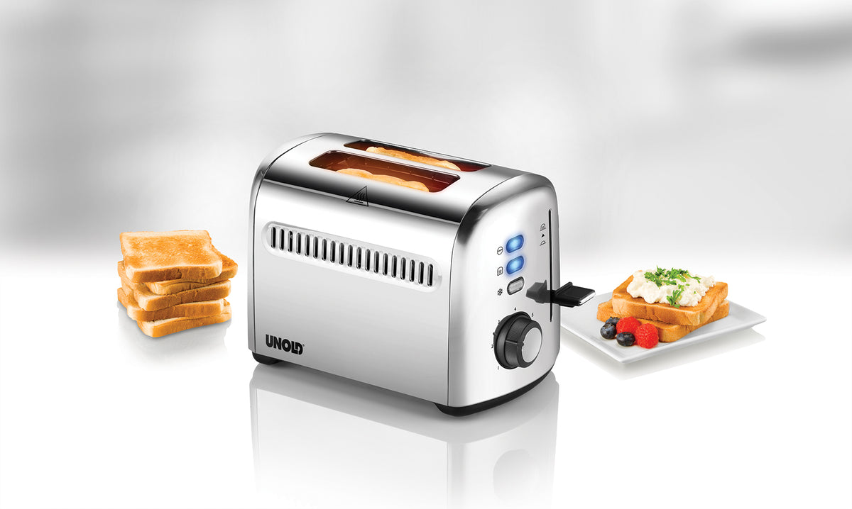 Der Unold Toaster 2er Retro in Silber steht auf einer weißen Fläche. Darin befinden sich zwei Brotscheiben. Daneben liegen gestapelte Brotscheiben und ein Teller Toast mit Gemüse und Obst.