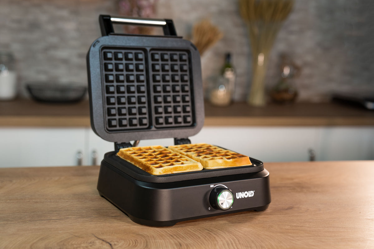 Ein schwarzes Waffeleisen mit geöffnetem Deckel zeigt zwei frisch gebackene quadratische Waffeln auf einer hölzernen Küchentheke an, mit einem verschwommenen Hintergrund aus Küchenutensilien und Dekor.