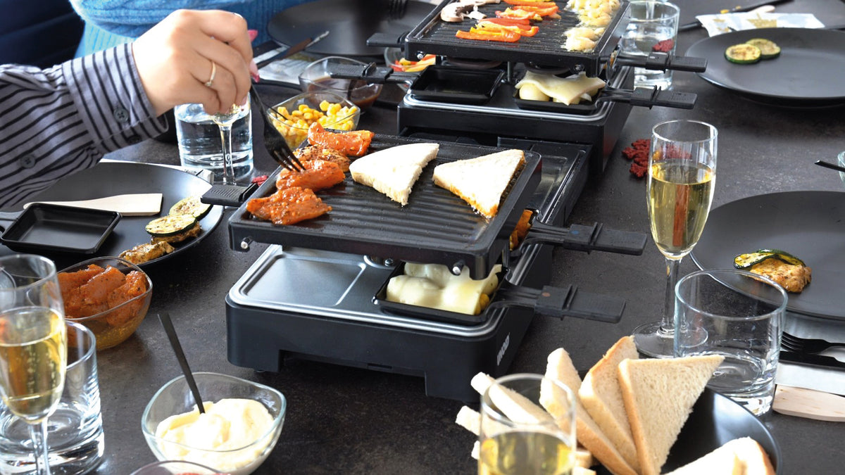 Menschen versammeln sich um einen Tisch und bereiten mit dem ausziehbaren Raclette-Grill von Unold Brot, Gemüse und Käse zu. Getränke, Teller und Besteck stehen auf dem Tisch, während eine Person das Essen auf dem Grill mit einer Gabel wendet.