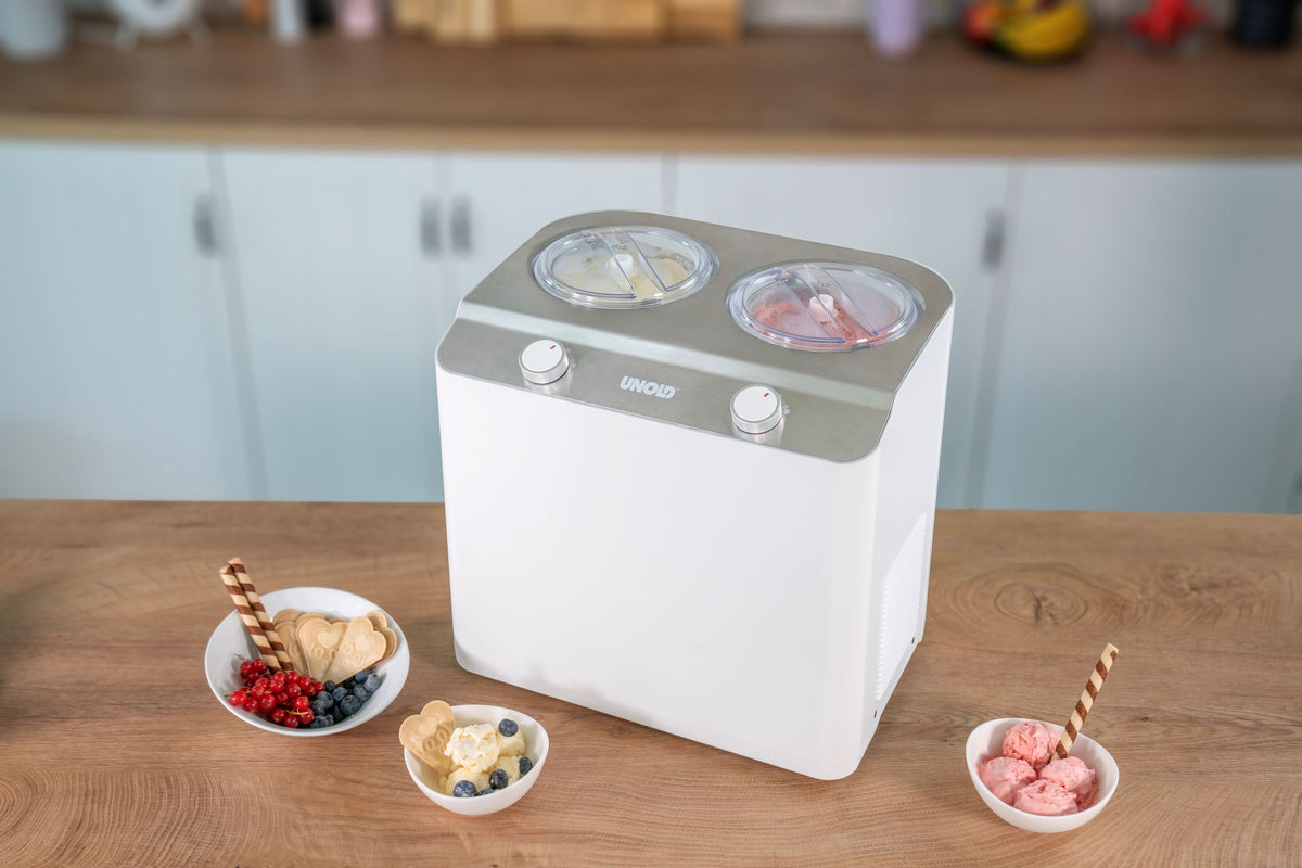Die Unold EISMASCHINE Doppio 2,4 L, eine weiße Eismaschine mit zwei Schüsseln, steht auf einer hölzernen Küchentheke und hat zwei Schüsseln darauf. Schüsseln mit Eiscreme und frischem Obst umgeben die Maschine; dahinter sind Küchenschränke und Küchenutensilien sichtbar.