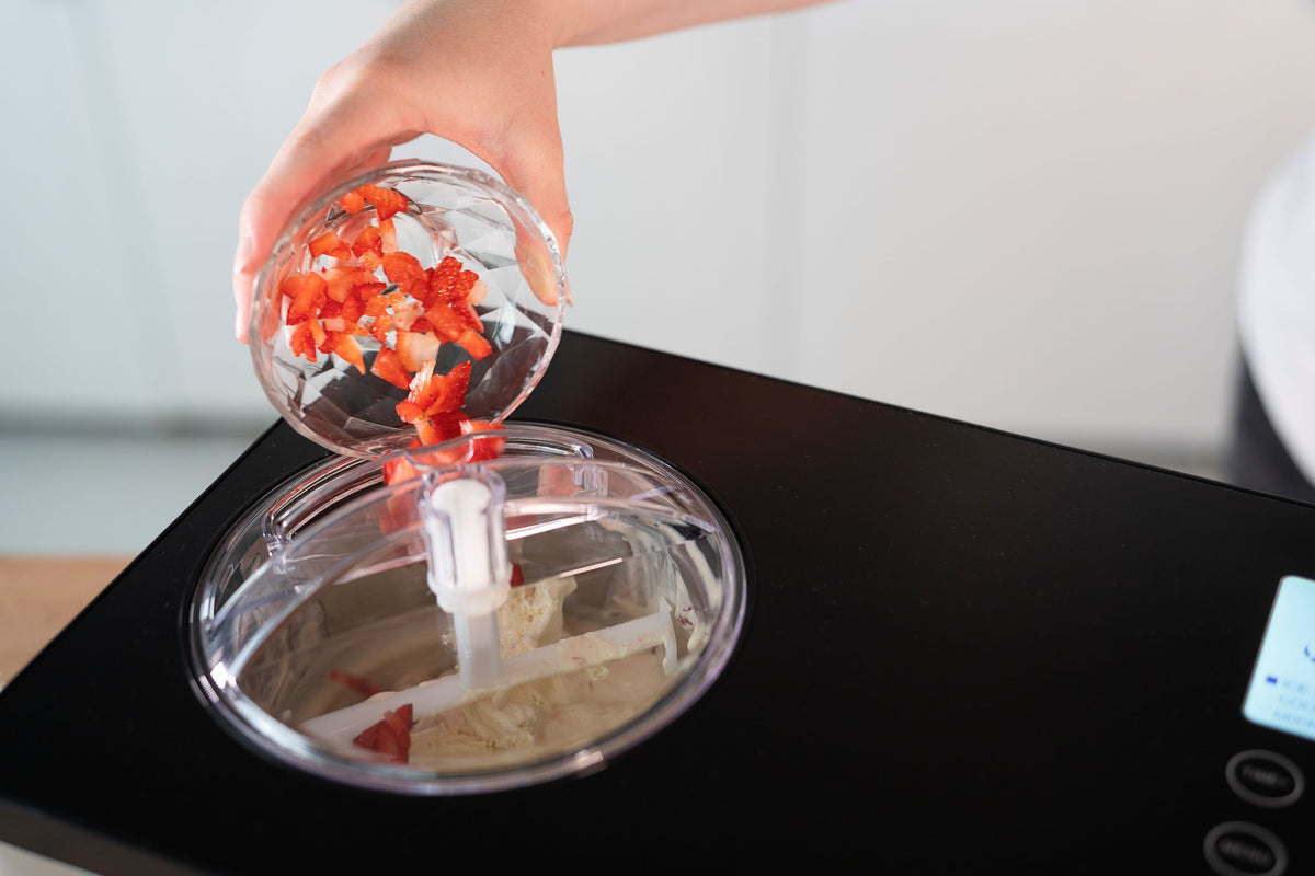 Eine Hand schüttet gehackte Erdbeeren aus einer kleinen Glasschale in die Unold Eismaschine Gusto 2 L, die teilweise gemischtes Eis enthält.