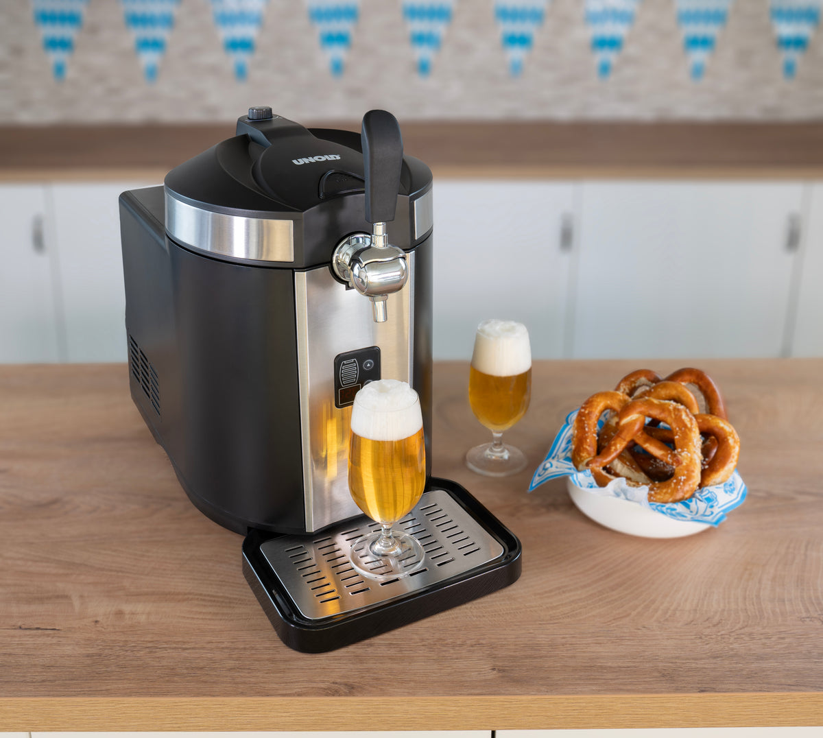 Eine Bierzapfanlage gießt Bier in ein Glas auf einer Holztheke. Daneben stehen zwei gefüllte Biergläser und eine Schüssel mit Brezeln. Im Hintergrund hängen blau-weiße Weihnachtsdekorationen.