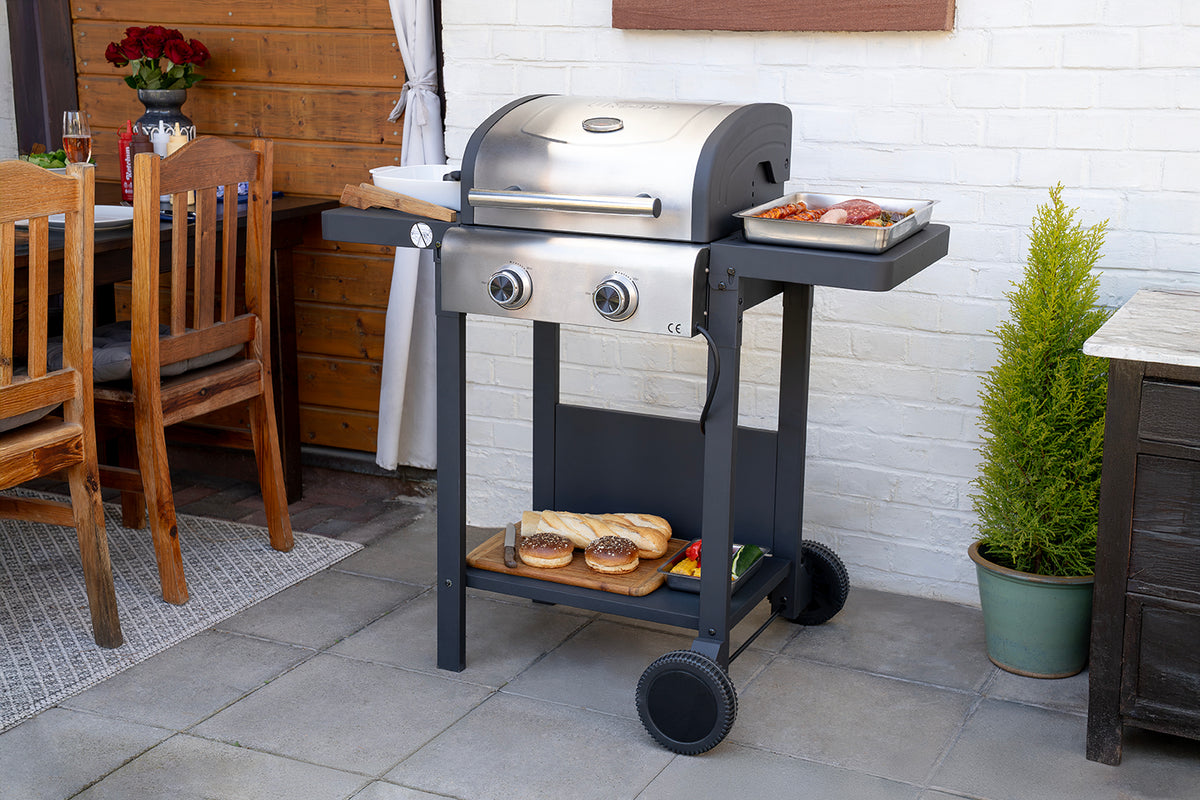 Der Unold Barbecue Grill-Wagen Buddy in Silber steht auf einer Terrasse, auf der Steaks und Würstchen gebraten werden, in der Nähe stehen ein Tisch und Stühle, auf der unteren Ablage liegen belegte Brötchen und Gemüse, und im Hintergrund stehen Grünpflanzen und eine Holzwand.