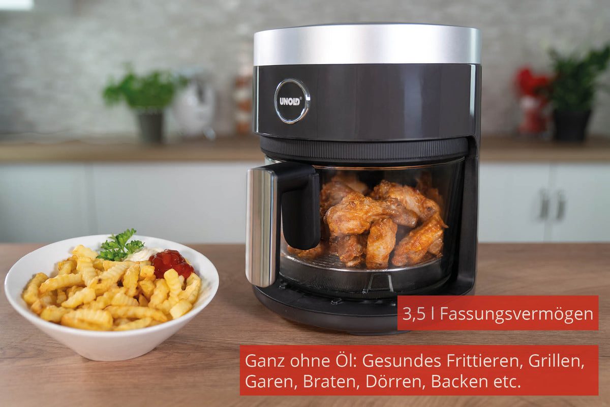 Die Unold Heißluftfritteuse Glas, eine schwarze Heißluftfritteuse mit 3,5 Litern Fassungsvermögen und ölfreiem Garen, steht mit gebratenen Hähnchenflügeln darin auf der Küchentheke. Davor liegen in einer weißen Schüssel Pommes Frites mit Ketchup und Petersilie.
