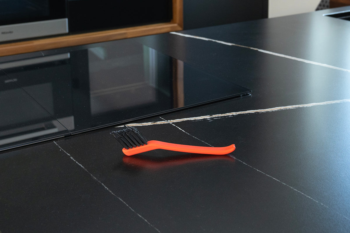 Die Unold ESGE-Zauberstab Reinigungsbürste, eine kompakte schwarz-orangefarbene Reinigungsbürste mit steifen Borsten, steht auf einer stilvollen schwarzen Arbeitsplatte mit weißer Maserung in der Nähe eines eingebauten Kochfelds in einer modernen Küche.