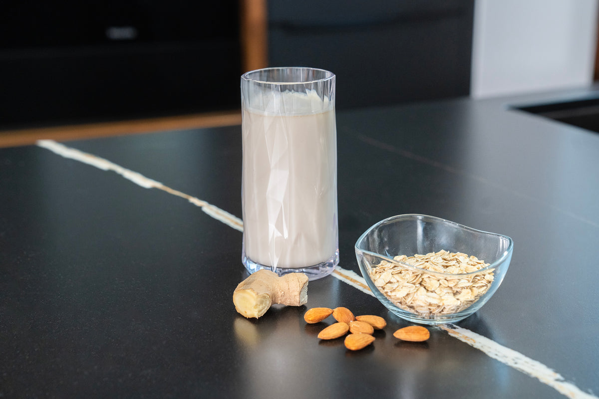 Auf einer dunklen Arbeitsplatte steht neben dem ESGE-Zauberstab Magic Fusion ein Glas Hafermilch, dazu eine kleine Schüssel Haferflocken, einige ganze Mandeln und ein Stück Ingwer.
