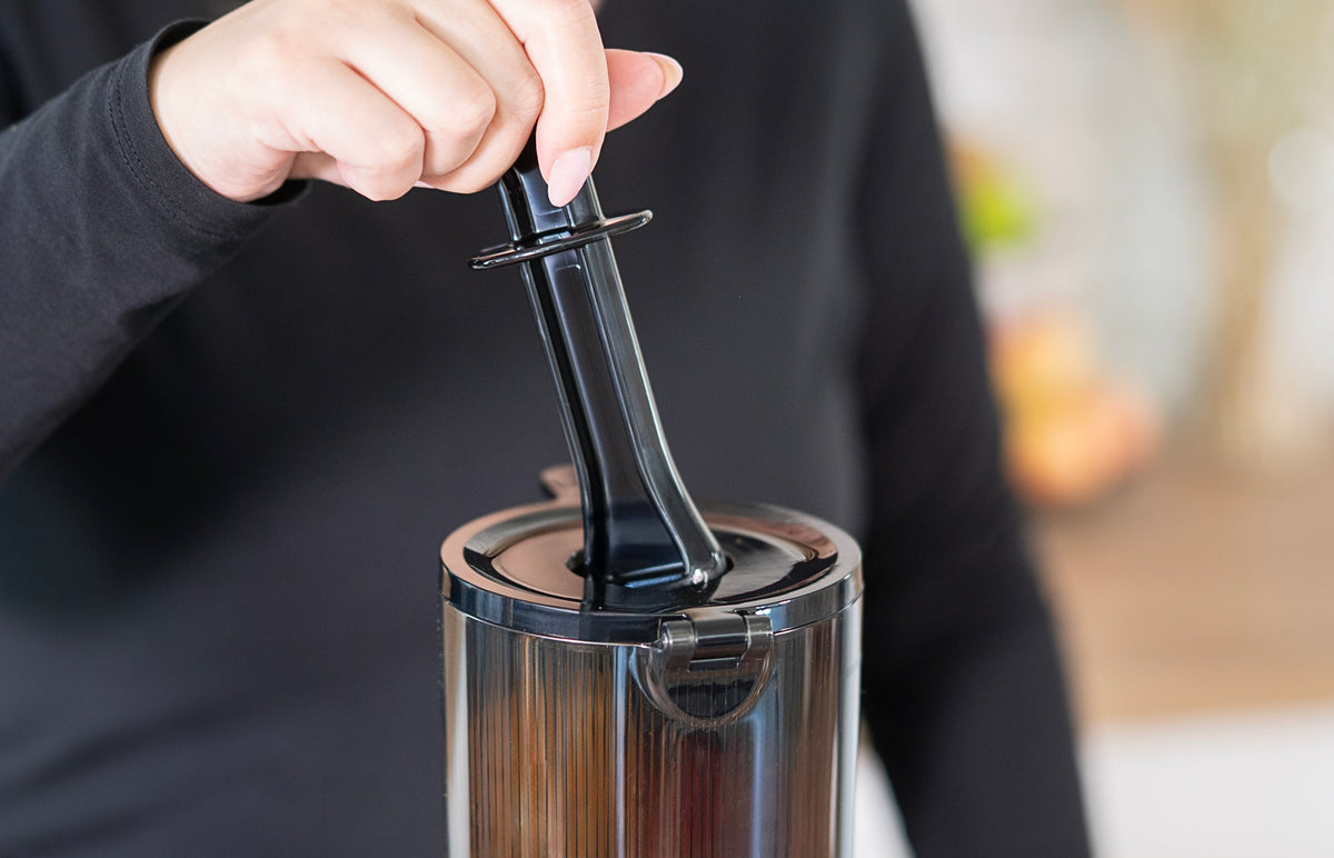 Eine Person in einem schwarzen Hemd drückt mit dem schwarzen Stampferwerkzeug Zutaten in den Einfüllschacht des Unold Slow Juicer Sam, vor einem leicht verschwommenen Hintergrund.