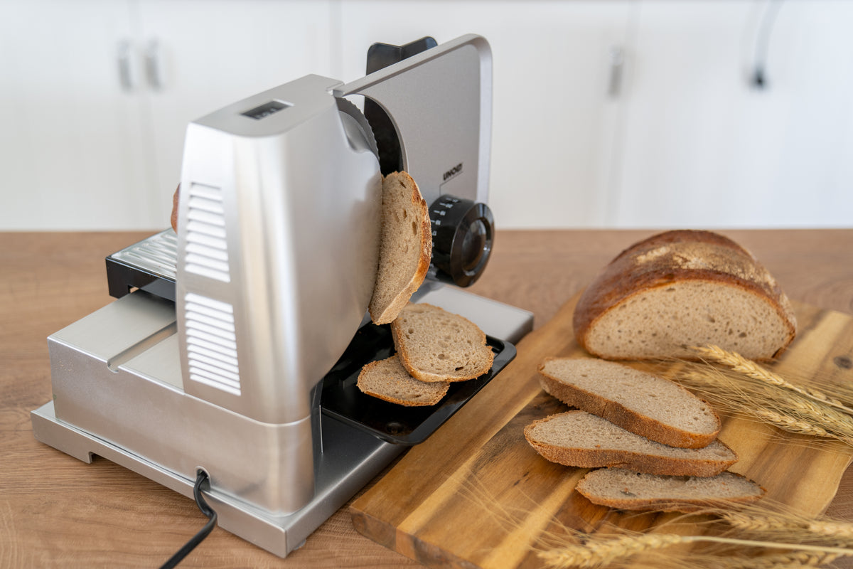 Die elektrische Brotschneidemaschine Unold Allesschneider Metall Plus in Silber schneidet einen Laib Schwarzbrot. Mehrere Brotscheiben liegen auf der Maschine, daneben ein Holzbrett mit Weizenhalmen vor einem hellen Küchenhintergrund.