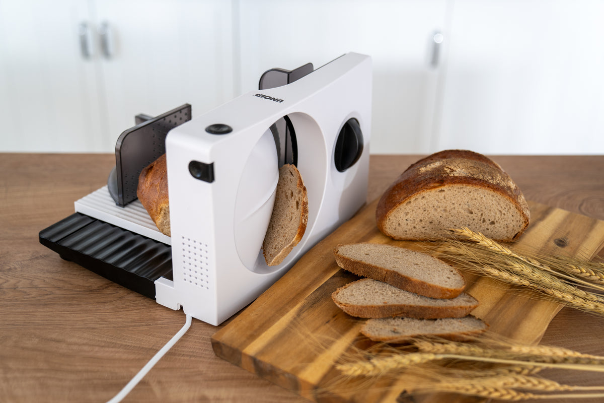 Der Unold Allesschneider Curve, ein elektrischer Weißbrotschneider, schneidet einen Laib Schwarzbrot auf einer Holztheke, während Weizenhalme und sauber geschnittene Scheiben auf einem Holzbrett daneben präsentiert werden.