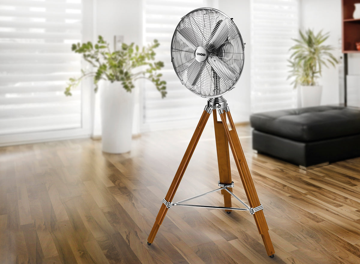 Der Unold STANDVENTILATOR KOLONIAL mit Metallkopf und dreibeinigen Holzbeinen steht auf einem polierten Holzboden in einem hellen Wohnzimmer mit großen Fenstern und Topfpflanzen.