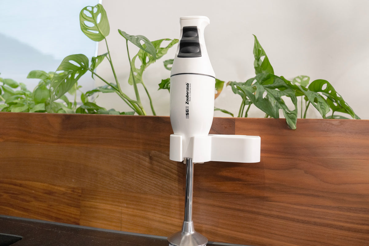 Der Unold ESGE-Zauberstab® M 100 Design Weiß ist an einer hölzernen Küchenwand montiert, mit grünen Blattpflanzen im Hintergrund.