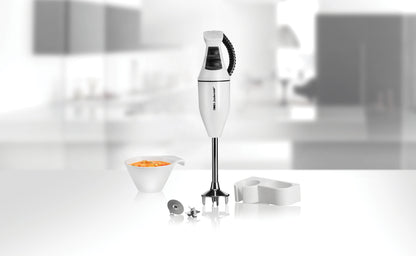 Der ESGE-Zauberstab 902105 Set E120 Select Stabmixer von ESGE Zauberstab steht aufrecht auf einer glänzenden Oberfläche neben einer Schüssel mit geschnittenem orangefarbenem Gemüse und verschiedenen Accessoires, im Hintergrund ist eine verschwommene moderne Küche zu sehen.