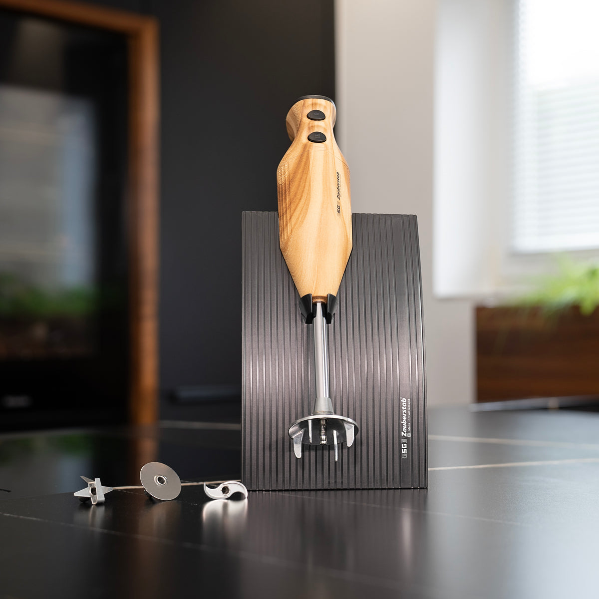 Der ESGE-Zauberstab Design Edition Douglasie Stabmixer von ESGE-Zauberstab steht aufrecht auf einem schwarzen gerillten Sockel mit drei Metallaufsätzen daneben auf einer modernen Küchentheke, mit einem Fenster und Pflanzen im Hintergrund.