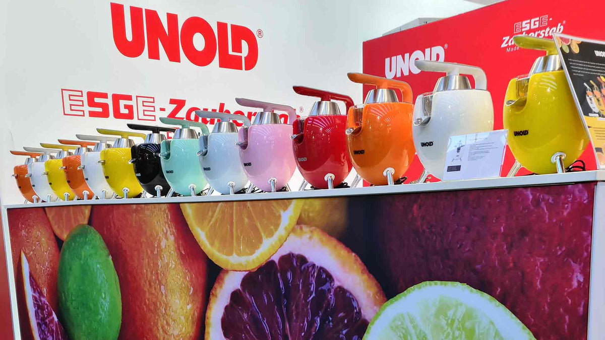 Auf einer Theke stehen mehrere bunte Mixer, jeder in einer anderen leuchtenden Farbe. Im Hintergrund sind ein großes UNOLD-Schild und Obstbilder zu sehen.