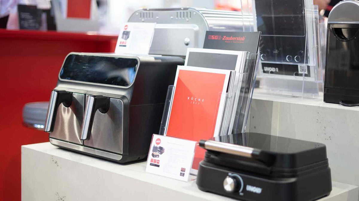 Eine Ausstellungstheke mit einer modernen Heißluftfritteuse aus Edelstahl, Broschüren in Haltern und einem schwarzen Elektrogrill, alles vor einem rot-weißen Hintergrund.