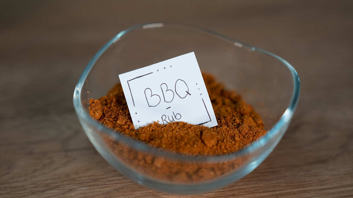 Eine Glasschale mit rotbrauner BBQ-Trockengewürzmischung steht auf einer Holzoberfläche. In der Schale befindet sich eine kleine weiße Karte mit der Aufschrift „BBQ Rub“.