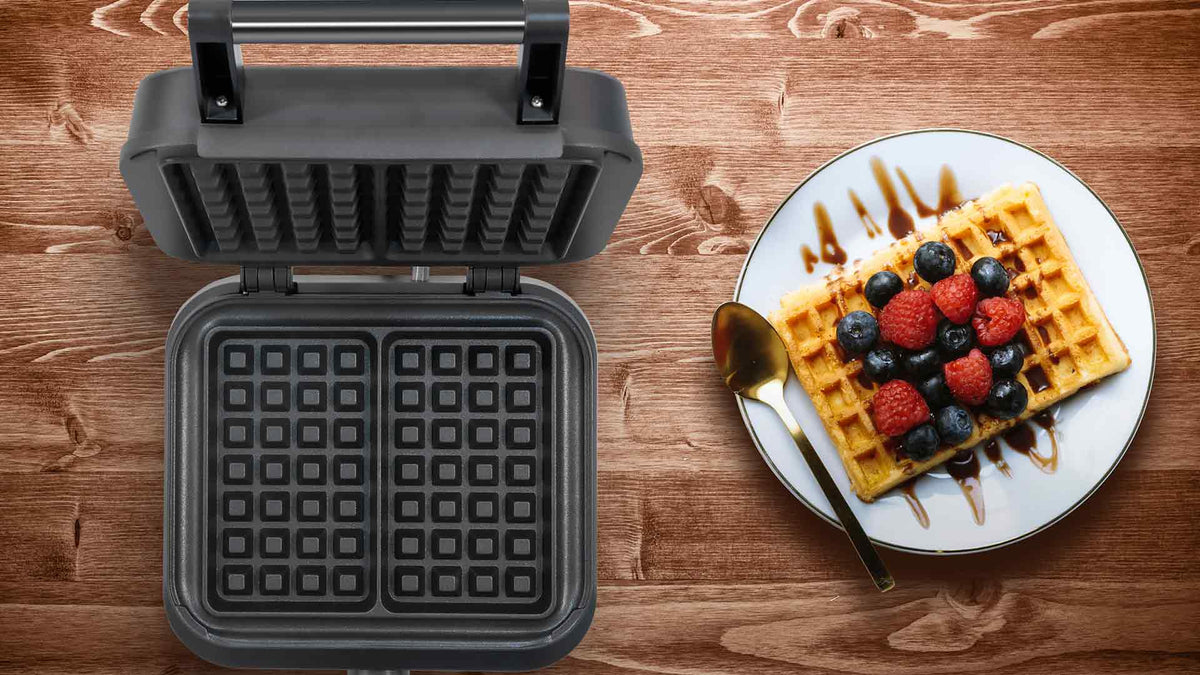 Auf einem Holztisch steht ein geöffnetes Waffeleisen neben einem Teller mit einer Waffel, die mit Blaubeeren, Himbeeren und Schokoladensoße garniert ist, und neben dem Teller liegt ein goldener Löffel.