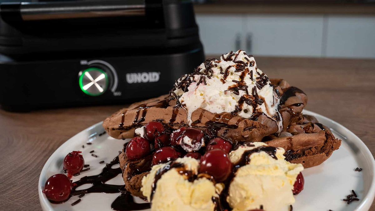Ein Teller mit Schokoladenwaffeln, garniert mit Schlagsahne, Schokoladensoße, Vanilleeis und roten Kirschen, auf einem Holztisch mit einem schwarzen Küchengerät im Hintergrund.