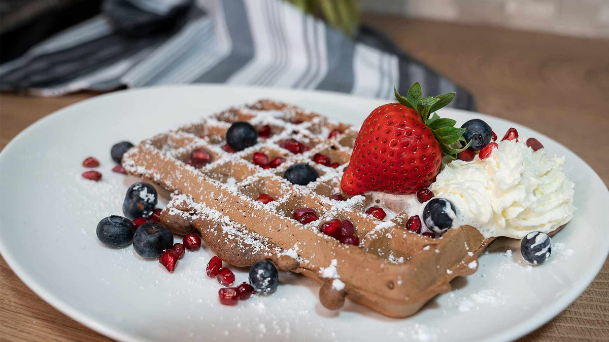 Eine Schokoladenwaffel mit Puderzucker, Blaubeeren, Granatapfelkernen, einer ganzen Erdbeere und einem Klecks Schlagsahne, serviert auf einem weißen Teller mit einer gestreiften Serviette im Hintergrund.