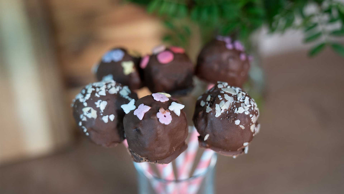 Eine Nahaufnahme von sechs Schokoladen-Cake-Pops auf gestreiften Stielen, dekoriert mit bunten Streuseln und essbaren Formen, die aufrecht in einem Glas stehen. Grünes Laub ist im Hintergrund unscharf.