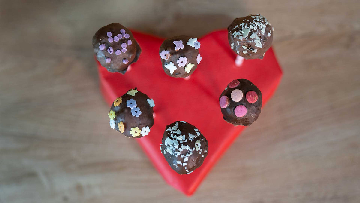 Fünf mit bunten Streuseln verzierte Schokoladen-Cake-Pops werden aufrecht auf einem roten, herzförmigen Ständer vor einem Hintergrund aus Holz präsentiert.