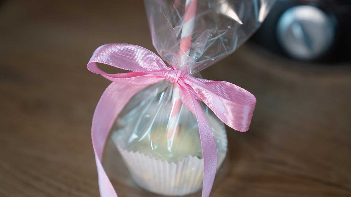 Ein in durchsichtige Plastikfolie eingewickelter Cupcake mit einer rosa Schleife, die um einen Stiel gebunden ist, liegt auf einer Holzoberfläche.