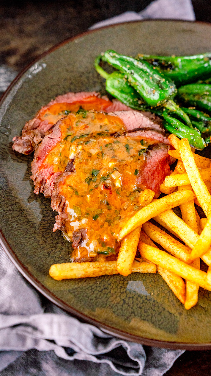 Ein Teller mit Steakscheiben, garniert mit Kräutersauce, goldbraunen Pommes Frites und sautierten grünen Paprikaschoten, serviert auf einer rustikalen Keramikschale.