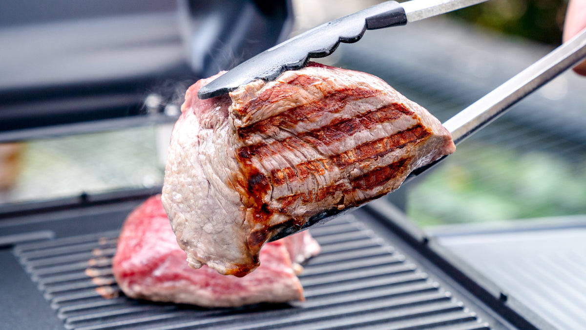 Nahaufnahme einer Metallzange, die ein gegrilltes Steak mit sichtbaren Grillstreifen über einem Grill hält, während im Hintergrund ein weiteres Stück Fleisch gart.