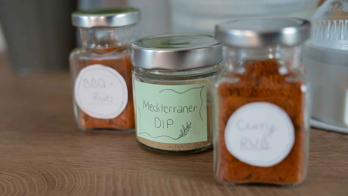 Auf einer Holzfläche stehen drei Glasbehälter mit Metalldeckeln, die mit den Aufschriften „BBQ Rub“, „Mediterraner Dip“ und „Curry Rub“ versehen sind und unterschiedliche Gewürzmischungen enthalten.
