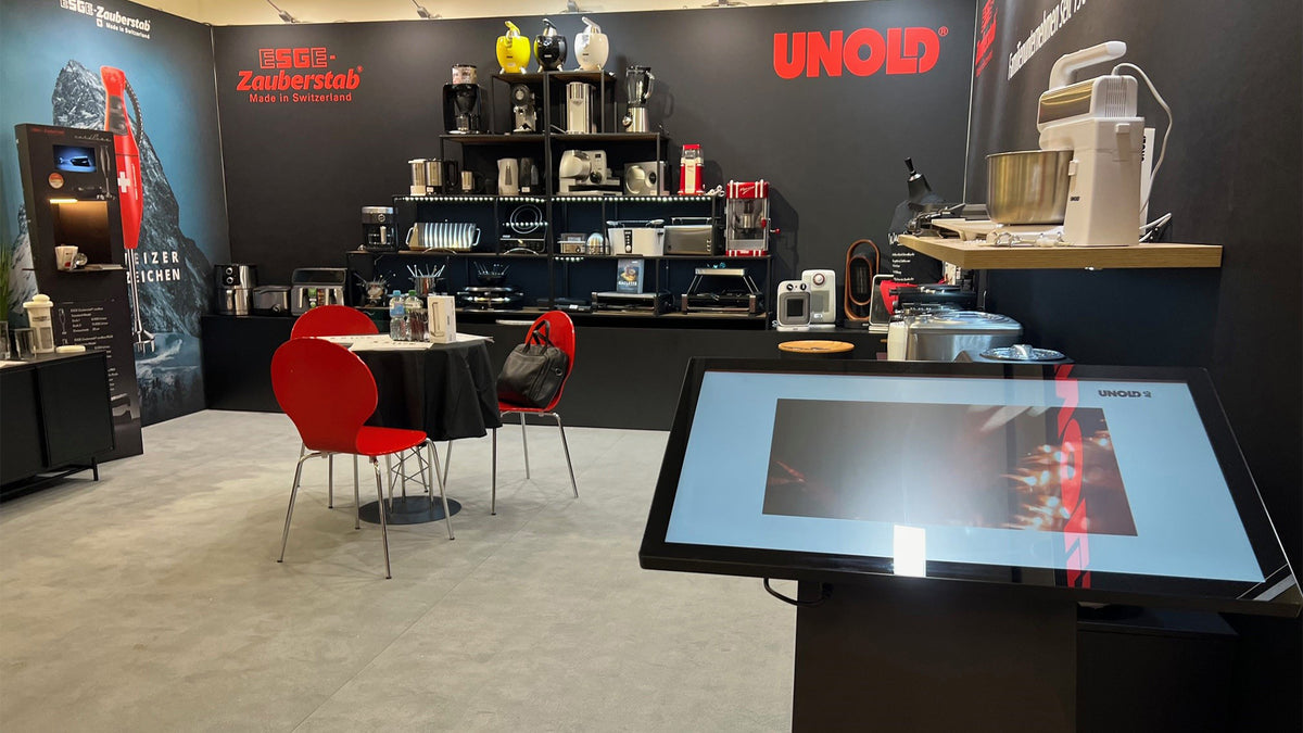Ein moderner Messestand präsentiert Küchengeräte auf Regalen und Theken, mit einem roten UNOLD-Logo an der Wand, einem Touchscreen-Display davor und zwei roten Stühlen um einen kleinen Tisch.