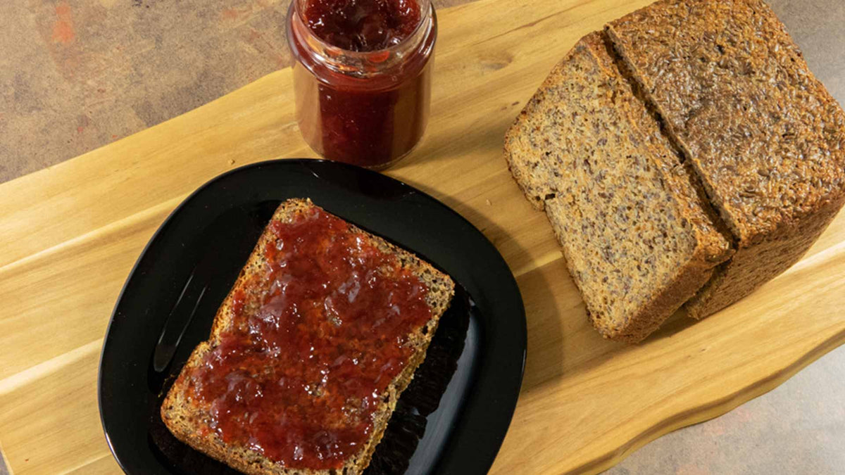 Eine Scheibe Mehrkornbrot mit roter Marmelade auf einem schwarzen Teller, ein Glas Marmelade und ein Laib Mehrkornbrot auf einem Holzschneidebrett.