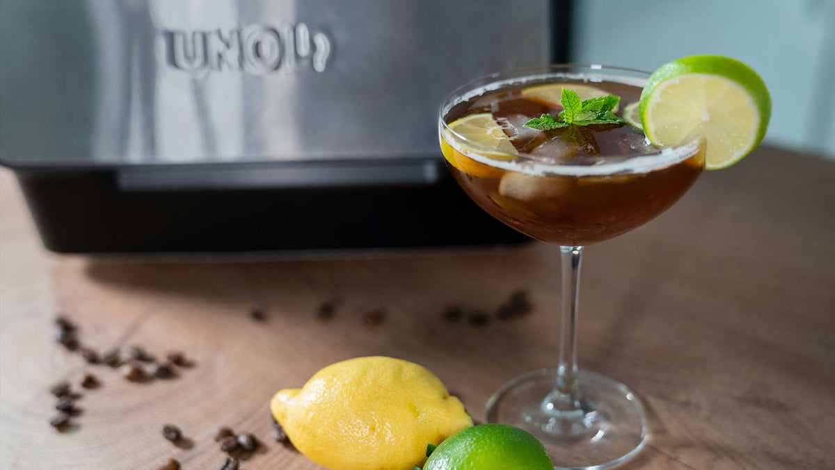 Ein Cocktailglas mit einem dunklen Eisgetränk, garniert mit einer Limettenscheibe und Minze, steht auf einem Holztisch. Daneben liegen eine Zitrone, eine Limette und verstreute Kaffeebohnen. Im Hintergrund ist ein Metallgerät zu sehen.