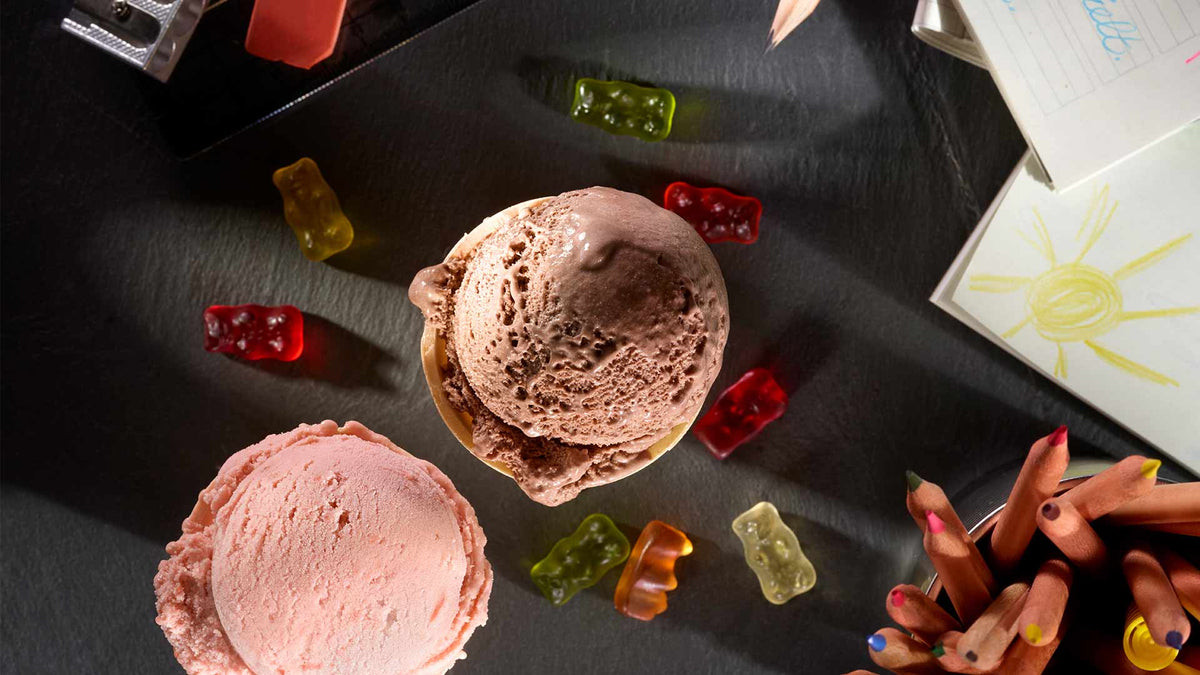Zwei Kugeln Eis, eine mit Schokolade und eine mit Erdbeere, sind umgeben von bunten Gummibärchen, Buntstiften und einer Kinderzeichnung mit einer Sonne darauf, alles angeordnet auf einer dunklen Oberfläche.