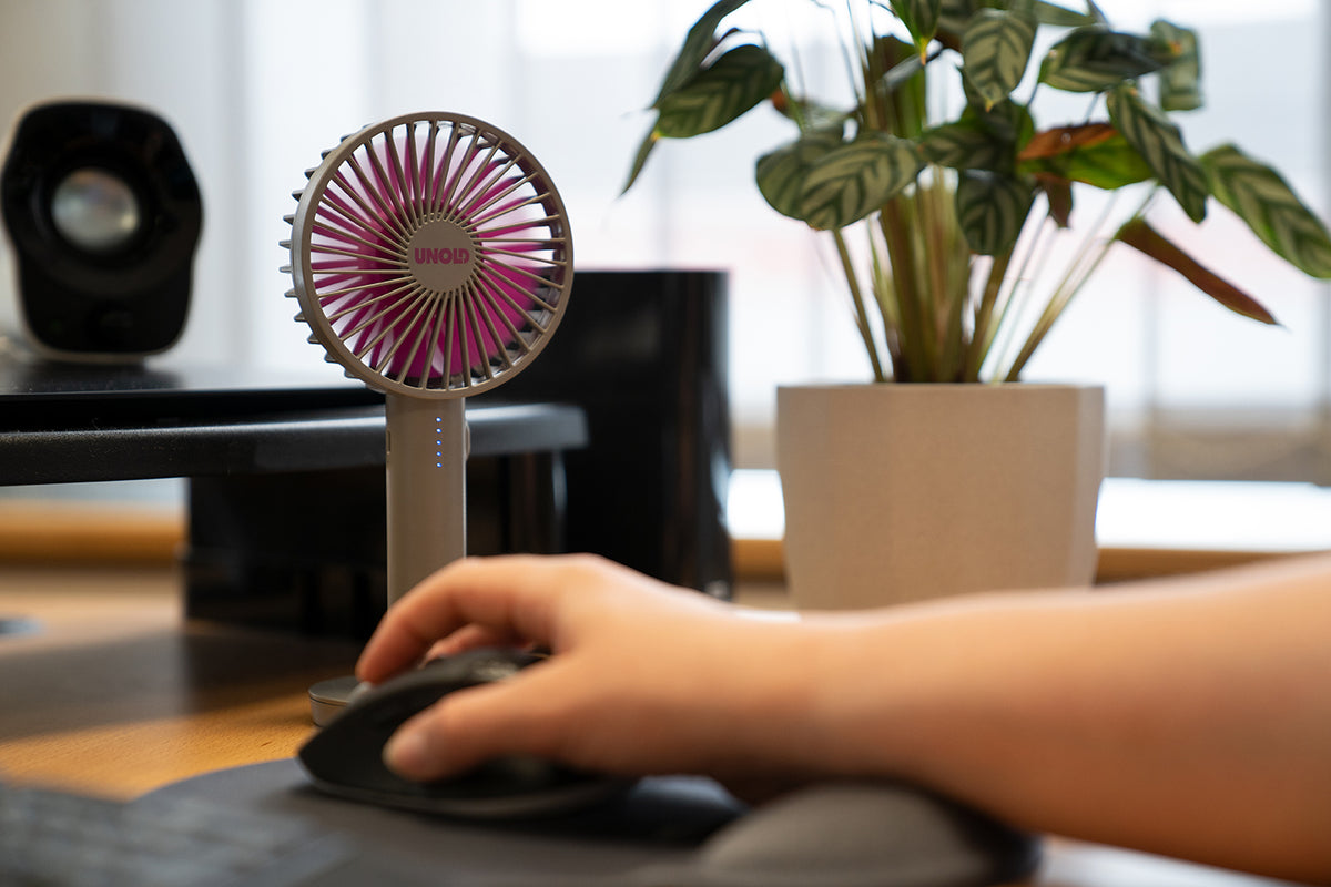 Eine Hand bedient eine Computermaus auf einem Schreibtisch neben dem Unold Handventilator Breezy II, einer Topfpflanze und Computerausrüstung, während im Hintergrund Sonnenlicht durch ein Fenster strömt.