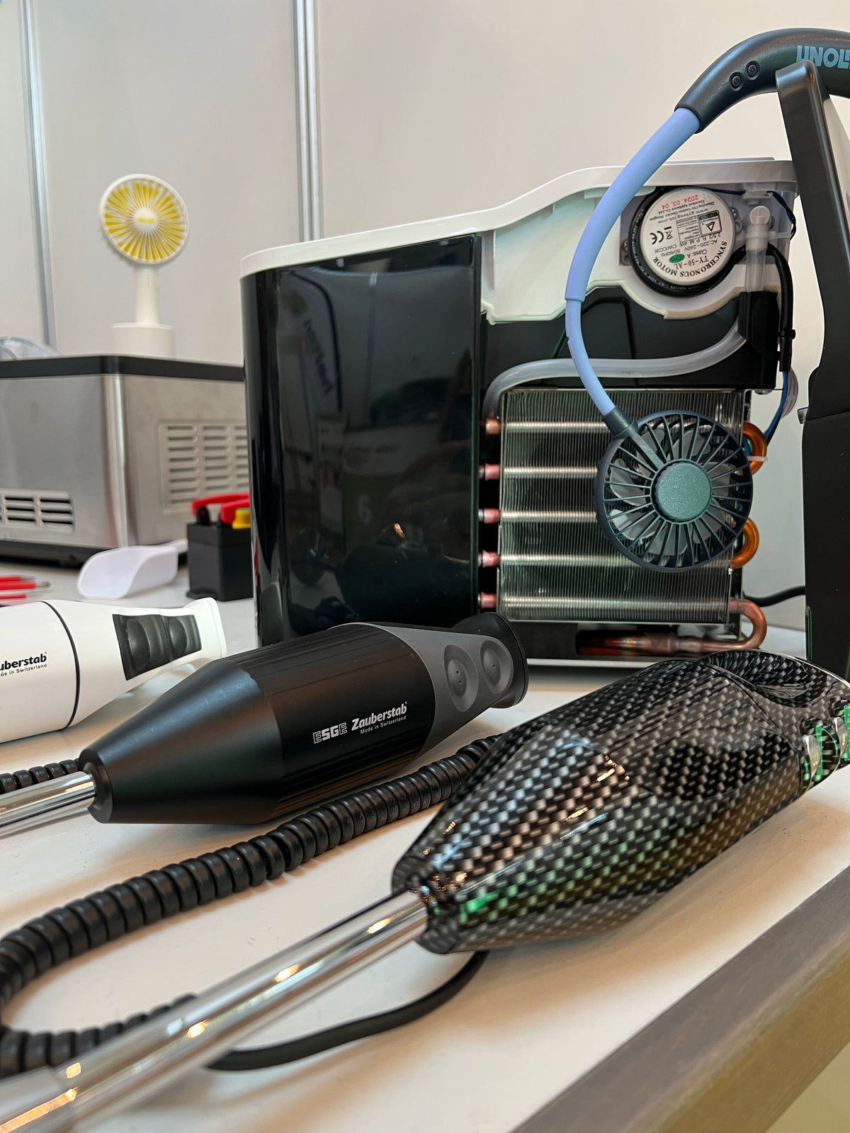 Nahaufnahme moderner Zahnarztgeräte auf einem Tisch, darunter ein Handgerät mit schwarzem Kohlefasermuster, ein schwarzes Gerät mit der Aufschrift „Zephyrtech“ und eine Maschine mit einem Kühlventilator und Spiralkabeln im Hintergrund.