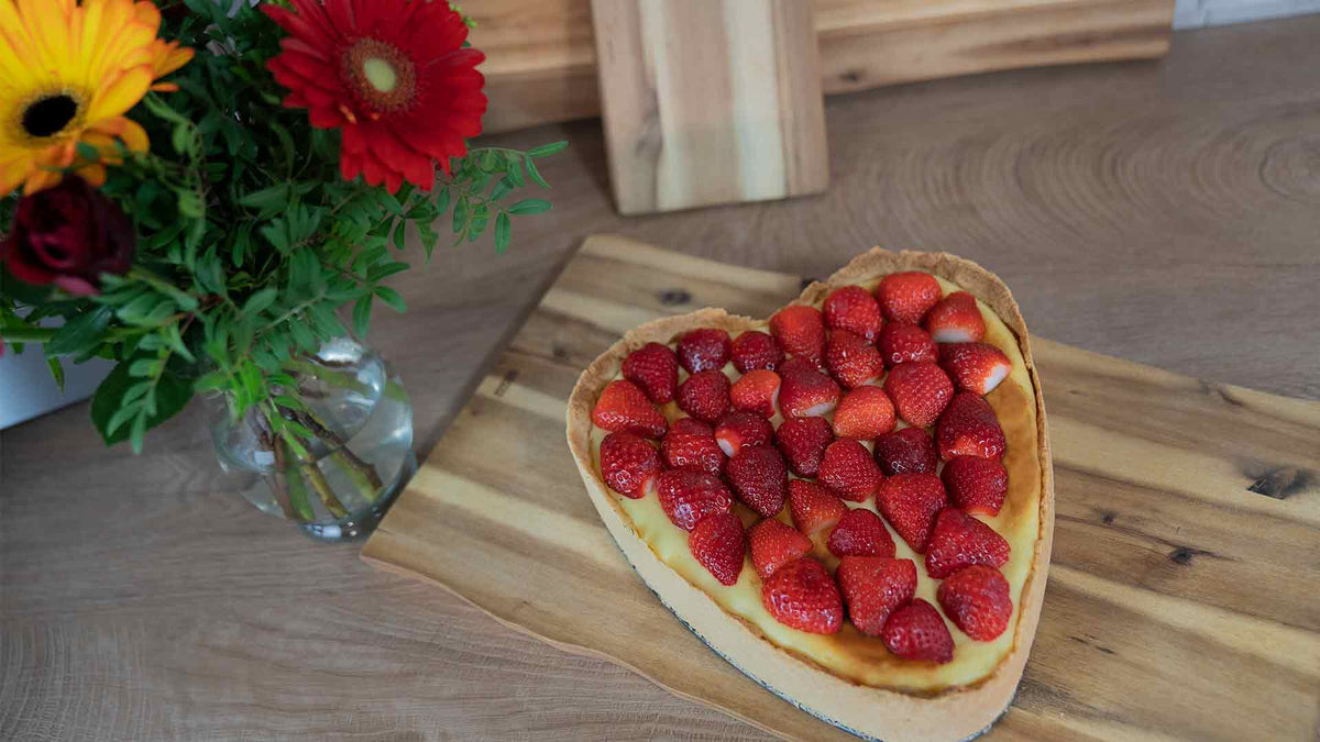 Ein herzförmiger Käsekuchen mit frischen Erdbeeren steht auf einem Holzschneidebrett neben einer Vase mit bunten Blumen auf einer Holzarbeitsplatte.