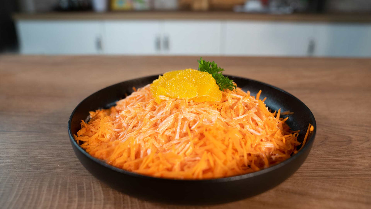 Eine schwarze Schüssel gefüllt mit geraspelten Karotten, garniert mit Orangenscheiben und einem Zweig Petersilie, steht auf einem Holztisch vor einem unscharfen Küchenhintergrund.