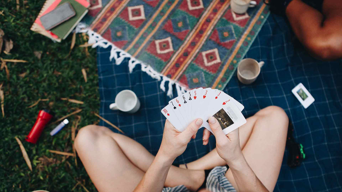 Eine Person sitzt im Schneidersitz auf einer Picknickdecke im Freien und hält einen Fächer mit Spielkarten in der Hand. Tassen, eine Taschenlampe, eine Kamera und ein Notizbuch liegen verstreut auf gemusterten Decken in der Nähe.