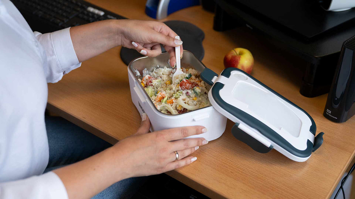 Eine Person sitzt an einem Schreibtisch und isst mit einer Gabel Reis und Gemüse aus einer weißen Lunchbox. Eine Computertastatur und ein Apfel liegen daneben auf dem Schreibtisch.
