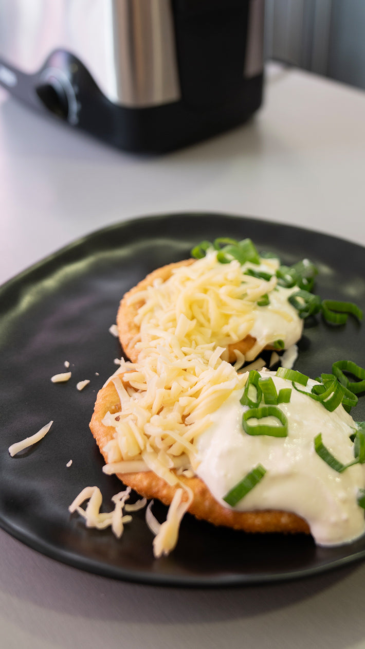 Auf einem schwarzen Teller liegen zwei Scheiben gebratenes Brot mit Sauerrahm, geriebenem Käse und gehackten Frühlingszwiebeln. Im Hintergrund ist ein Küchengerät unscharf zu sehen.