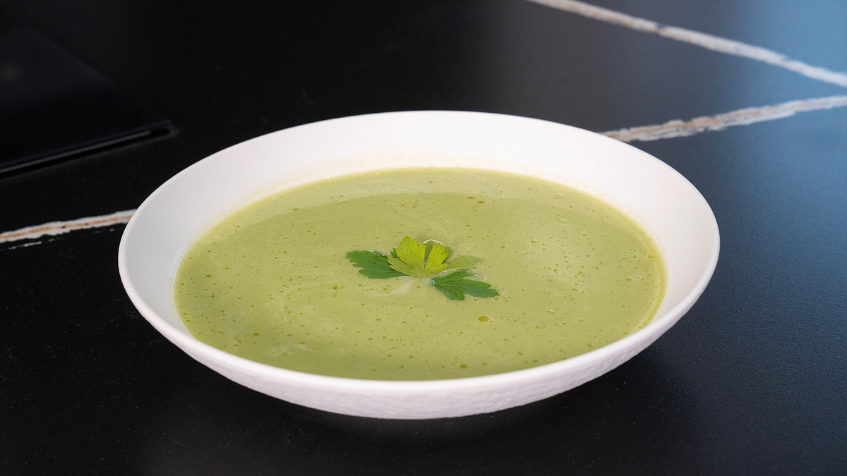Eine weiße Schüssel gefüllt mit cremiger grüner Suppe, garniert mit einem Zweig frischer Petersilie, steht auf einer schwarzen Oberfläche mit hellen Streifen im Hintergrund.