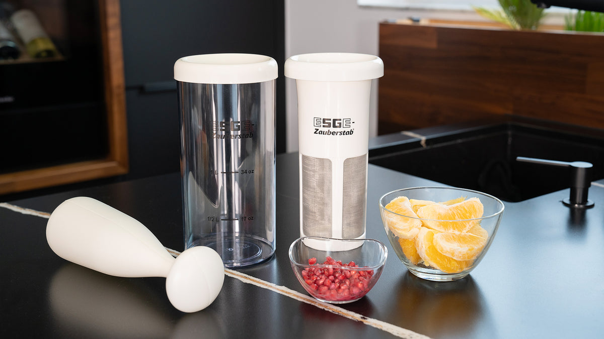 Auf einer Küchentheke steht ein weißer Stabmixer mit zwei Mixbehältern neben einer Glasschale mit Granatapfelkernen und einer Schale mit Orangenscheiben. Die Behälter tragen die Aufschrift „ESGE-Zauberstab“.