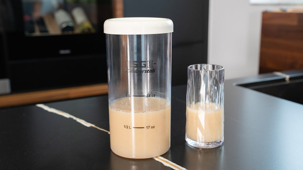 Ein hoher transparenter Shaker mit weißem Deckel, teilweise gefüllt mit einer beigen Flüssigkeit, steht neben einem Glas mit dem gleichen Getränk auf einer schwarzen Küchentheke.