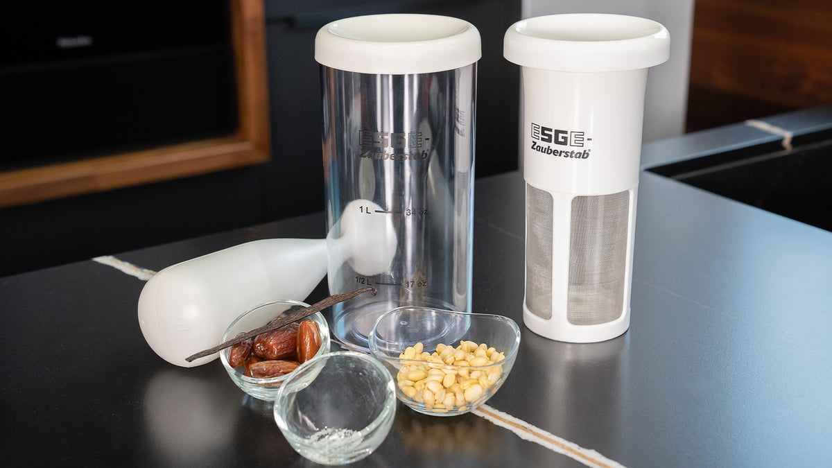 Zwei durchsichtige Plastikbehälter mit weißen Deckeln, ein weißer Stabmixer, Datteln, Vanille, Pinienkerne und eine kleine Glasschale, angeordnet auf einer schwarzen Arbeitsplatte.