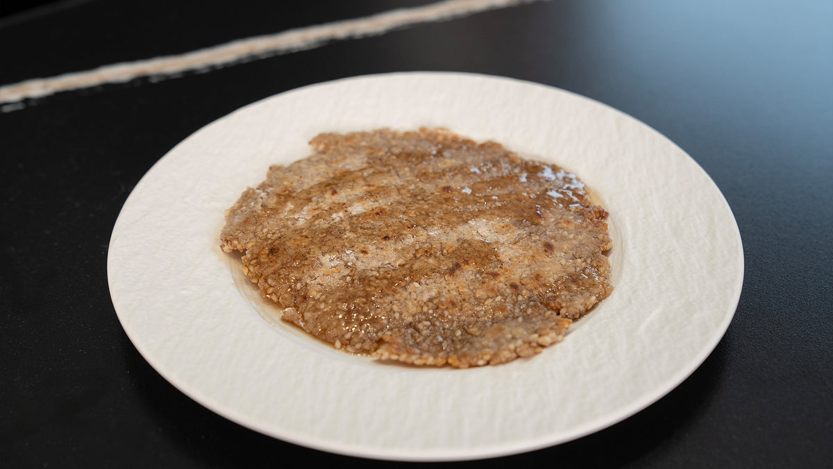 Ein weißer Teller mit einer dünnen, ungleichmäßigen Schicht brauner, körniger Paste auf einer dunklen Oberfläche. Die Paste wirkt feucht und hat eine leicht glänzende Textur.