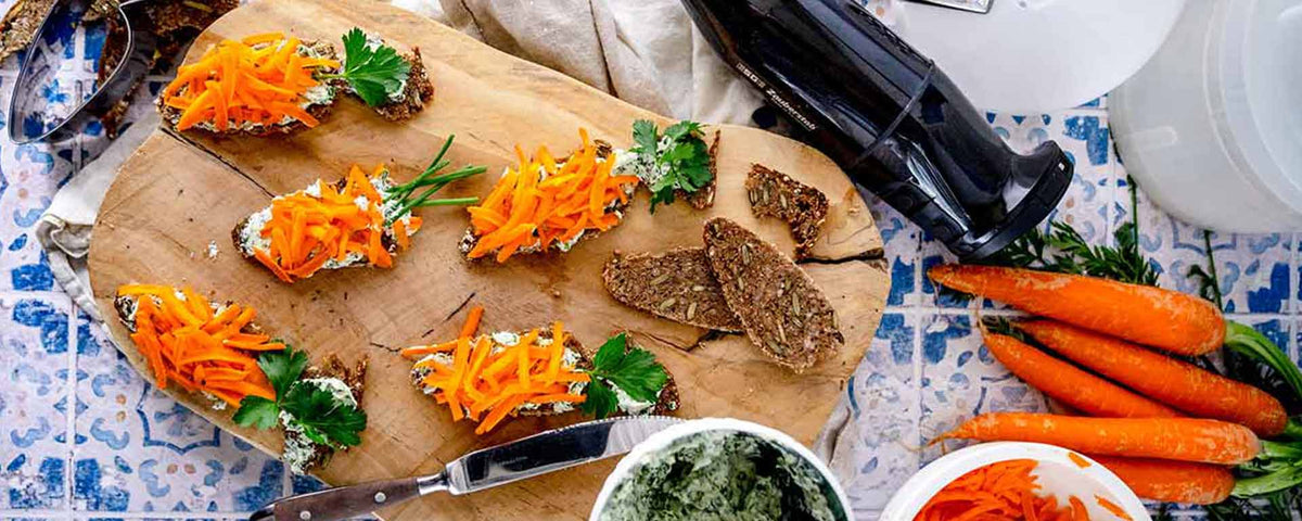 Auf einem gefliesten Tisch liegt ein Holzbrett mit Brotscheiben, belegt mit Frischkäse, geraspelten Karotten und frischen Kräutern. Daneben liegen ganze Karotten, ein Messer, eine Küchenmaschine und eine Schüssel mit grünem Brotaufstrich.