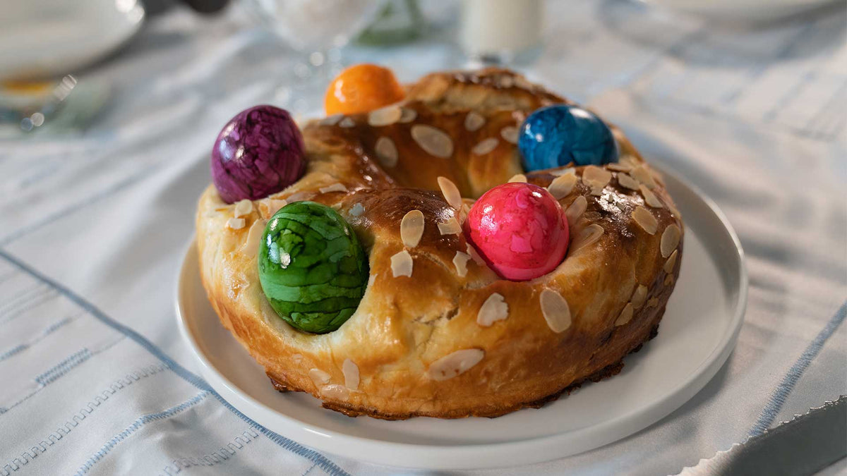 Auf einem weißen Teller auf einer hellblau-weißen Tischdecke liegt ein rundes süßes Brot, das mit Mandelblättchen belegt und mit bunten, marmorierten Ostereiern dekoriert ist.