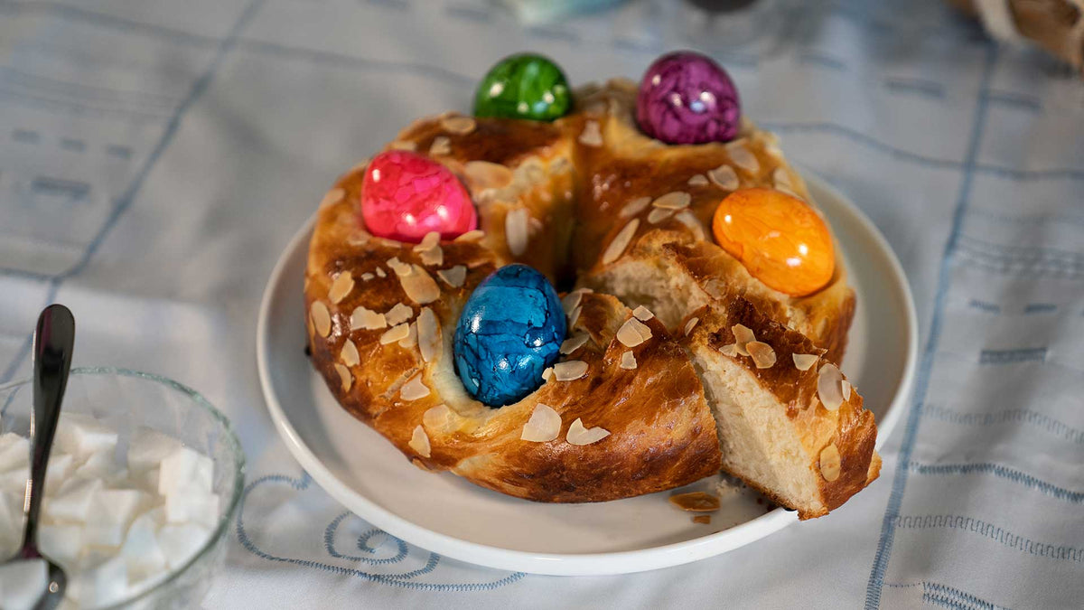 Ein rundes süßes Brot mit Mandelblättchen und bunt gefärbten Eiern liegt auf einem weißen Teller auf einem Tisch mit gemusterter Tischdecke.