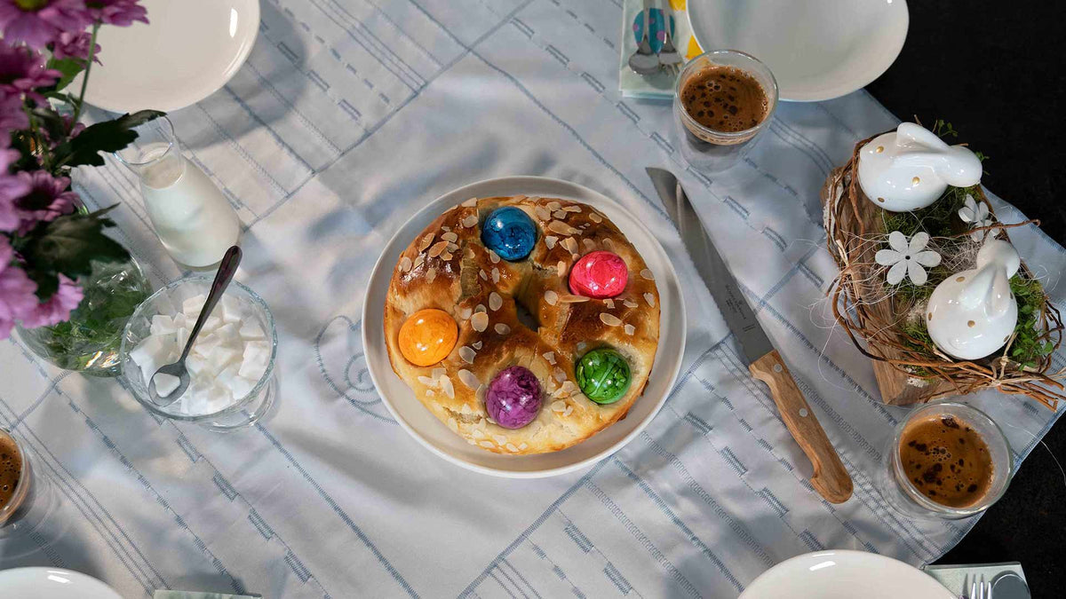 Ein gedeckter Kaffeetisch mit einem süßen Brot, das mit bunten Ostereiern und Mandeln belegt ist, umgeben von Kaffeetassen, Blumen, Schlagsahne, Milch und Keramikhasen als Dekoration.